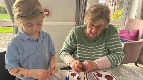 Lili wearing a blue school uniform short sleeved top sitting at a table with Shirley Robinson, wearing a light green top with white stripes. They are both looking down at the table which has cards which they are decorating