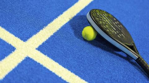A padel bat with a tennis ball on a blue and white court.