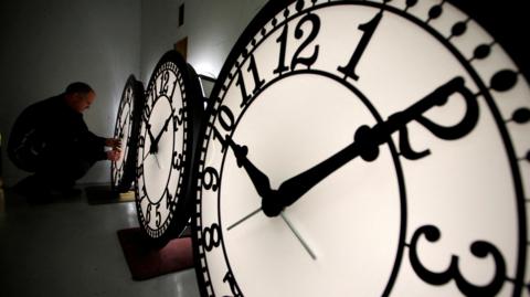 Three very large black and white clock faces, lit from behind. They are all set at nine minutes past ten. A man bends down as he fiddles with the hands of one clock.