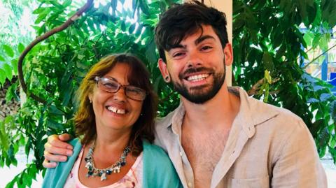 Olli and Sarah are together and smiling. He has his arm around her and is wearing an open beige shirt. She is wearing glasses and a green cardigan over a top.