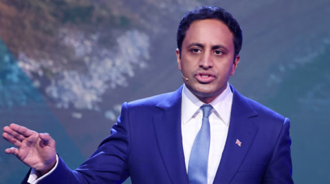 Reform UK home affairs spokesman Zia Yusuf speaks during a press conference in London. He wears a royal blue jacket with a white tie and a paler blue tie. He has short dark hair and is wearing a small union flag badge.