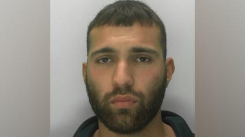 A police custody mugshot of Aaron Akram. He has short dark and facial hair. He is looking directly at the camera with a blank expression.