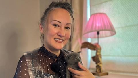 Frankie, a white woman with dark brown hair tied up in a bun smiles at the camera. She has well done make up with neatly stenciled eyebrows, glamorous. In her hands she holds a small grey kitten that is licking the tip of her thumb.