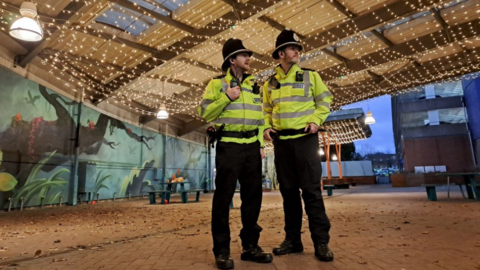 Two police officers in yellow jackets standing in an empty warehouse unit with christmas lights overhead