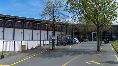 A view of the entrance of Beau Sejour. It is a building clad in white and glass. The road leading up to the centre has disabled parking and trees. 