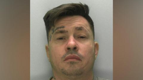 A mugshot of a man in his late 30s with quite a wrinkly forehead and a tattoo of a crown and the name "Victoria" above his left eye. He has brown hair swept over to one side. He is wearing a grey sweatshirt and standing in front of a light grey or white wall. He looks uncomfortable.