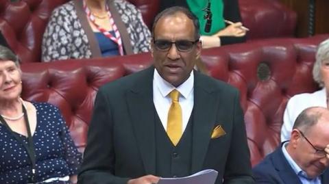 A man stands in a row of red leather seats. He is wearing glasses, a dark suit and yellow tie and is holding papers stapled together between his hands. The man next to him has his face in his hands while the two women in the row behind him look at him from either side.