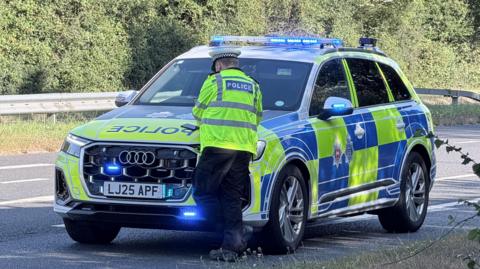 A police car with a police officer stood in front of it. The car is on a tarmac road surrounded by bushes.