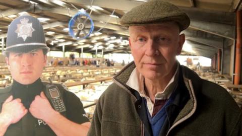 A man in a brown flat cap and brown jacket in a large barn with animals in the background. There is a life-sized cut out of a police officer to his right hand side.