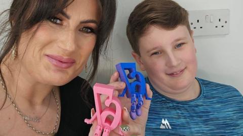 Karlla, a woman with dark hair, andAlfie, a pre-teen boy, sit side-by-side. They are each holding up small plastic statues of the number '67' with little arms and legs attached. The numbers appear in reverse, as in a mirror image, as they have taken a selfie.