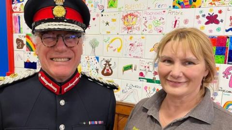 A closeup of a woman in a grey polo and a man in a black military uniform  standing by a wall with children's drawings all over it. 