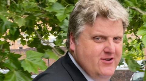 Gregory Hill, a man in a black suit jacket, white shirt and striped tie. He has blonde hair and is pictured outside court in Norwich.