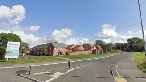 A Google Streetview looking north east from the Welford Road roundabout in Wigston. Part of a housing development surrounded by a green space and a road can be seen.