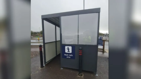 Grey multi faith area that is shaped similar to a bus stop in Bristol Airport's car park on grey day.