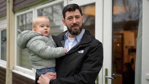 Tom standing outside his flat on a cold winter's day holding his little boy in his arms