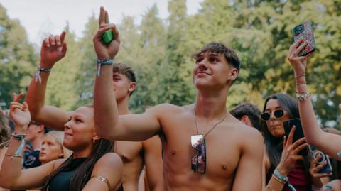 Young men and women raising their right hands while watching a band, with trees behind them