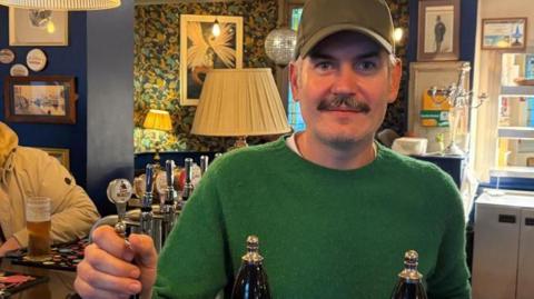 A man in a green jumper rests his hand on a bar pump in a pub. He looks directly at the camera and wears a khaki baseball cap and has a moustache.