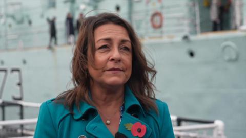 Kent County Council leader Linden Kemkaran is interviewed by the BBC, wearing a turquoise overcoat with a memorial poppy in the lapel, standing in front of a boat at the Historic Dockyard in Chatham.