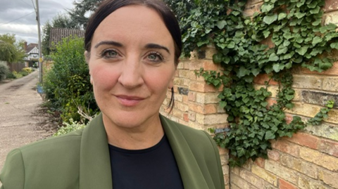 Kath Sansom, founder of the patient-led campaign group Sling the Mesh, stood outside next to a brick wall. She is looking directly into the camera and has on a green blazer jacket over a black top. The wall behind her is covered in green ivy. 