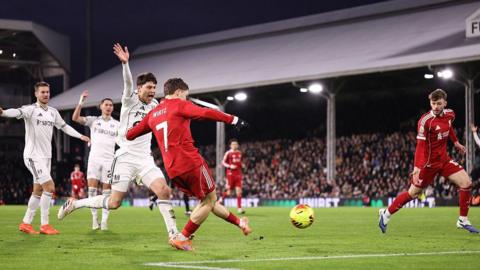 Liverpool's Florian Wirtz scores against Fulham. He is in the penalty area and has struck a yellow ball with his left foot. Liverpool's kit is red and he is wearing orange boots. A Fulham player, wearing all white, is next to him and is raising his right arm to call for offside. Several other players can be seen in the background along with thousands of fans in one of the stands.