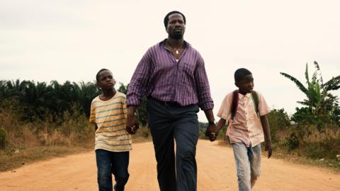 A man in a purple shirt walks down a sandy road holding hands with two young boys