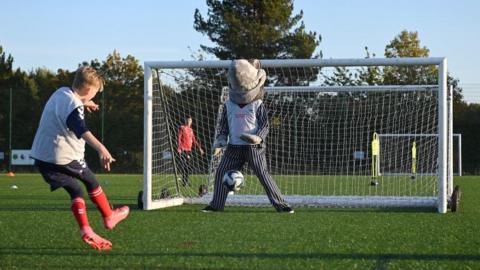A boy dressed in a football kit has just kicked a football into a goal, in a penalty shot against a shark mascot that stands in the middle of a football goal. This is taking place on an artificial grass pitch, with a blue sky in the background.