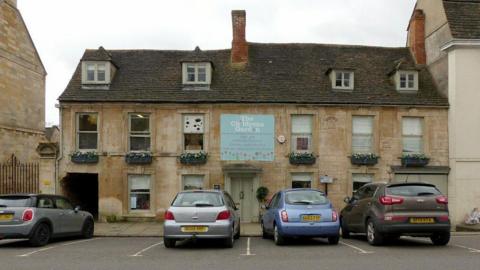 A sandstone building with a sign that reads "The Children's Garden Day Nursery". It has 4 windows in the slate roof. There are 6 windows on the first floor and 4 windows and a door on the ground floor. There is a passage through the building too. There are 4 cars parked in front in the parking spaces.