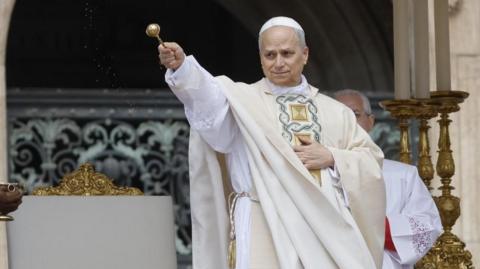 Pope Leo sprinkles holy water during his inauguration mass. he is wearing cream-coloured robes and holding a golden papal artefact.