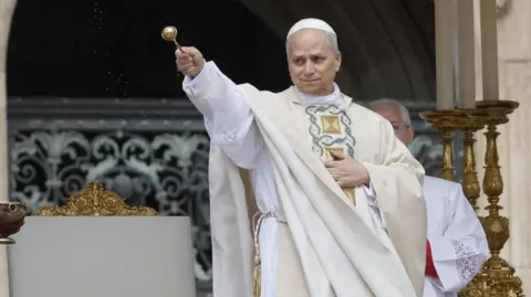Pope Leo sprinkles holy water during his inauguration mass. he is wearing cream-coloured robes and holding a golden papal artefact.