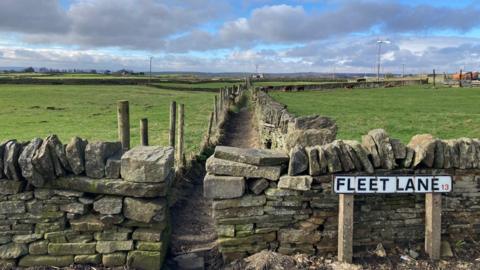 A road sign spelling out Fleet Lane in front of dry stone walls and a footpath between green fields 