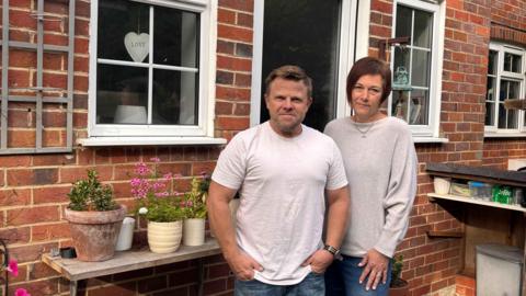 Two people stood outside the open door of a red brick house, looking cross. The house has white-framed windows and there is a ledge with flower pots on it beside them.