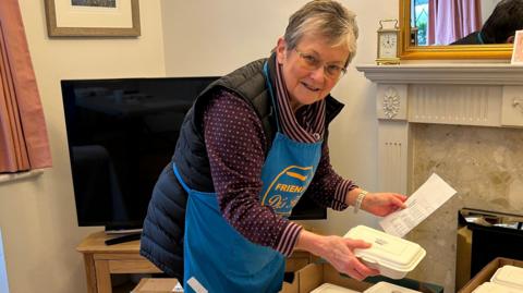 A woman putting white boxes into a larger box. She is wearing a blue apron and behind her is a television set and a shelf above a heater. The shelf has a clock on it and there is a mirror above it. 