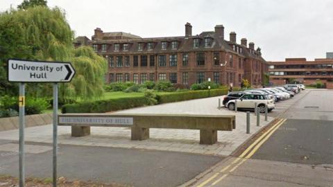 A Google street view of the main entrance to the University of Hull. A white sign reads 'University of Hull' and is pointing to the right. Behind is a car park and large red-brick building with black window frames.