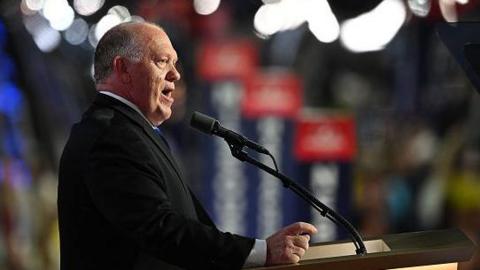 Tom Homan speaks into a microphone in front of a lectern. He wears a black suit jacket and white shirt.