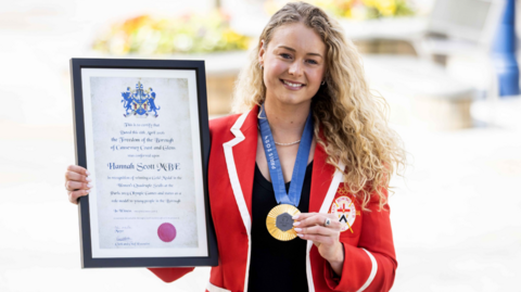 Hannah Scott is standing outside Coleraine Town Hall. She is wearing a red blazer with a white trim around the lapels and cuffs. She has blond curly hair and is holding a framed certificate of her freedom of the borough