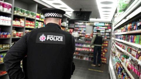 A police officer - seen from the back - entering a convenience store, with drinks in a fridge on the right and groceries in shelving on the left. He wears a black jacket with Greater Manchester Police written on the back and a police hat 