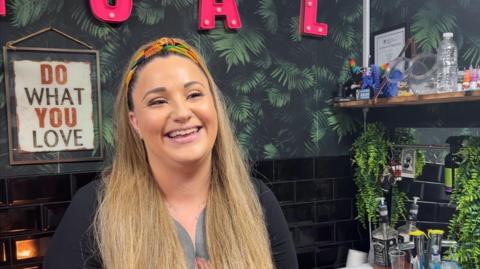 A smiling, heavily-tattooed young woman with long fair hair, tied back with a multi-coloured hairband in a colourful tattoo parlour.