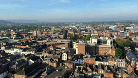 A view from the top of Gloucester Cathedral. Brown brick buildings can be seen below and hills in the background. 
