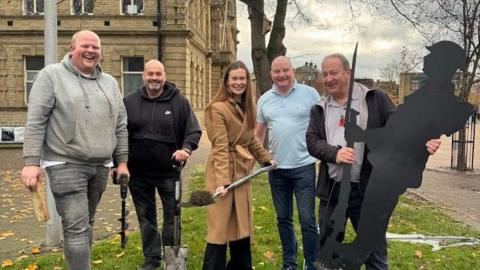 A group of five people standing on a grassy area near a hole in the ground, holding shovels and tools. One person is holding a black silhouette cutout of a soldier with a rifle.