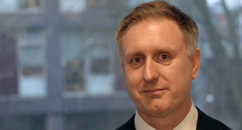 A man with short light brown hair wearing a shirt with a tie. He is looking into the camera. 