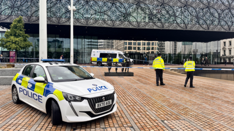 A police car parked on a paved area in front of a large building with a patterned metal front and a number of police officers in yellow jackets standing around
