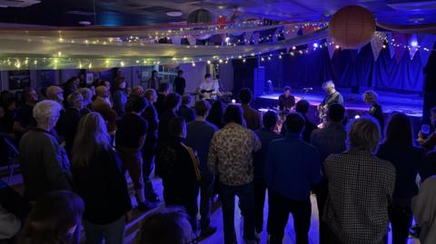 The inside of The Crescent on a gig night. There is a disco ball with fairy lights and bunting with a band playing in front of the stage. The audience face away from the camera towards the band.