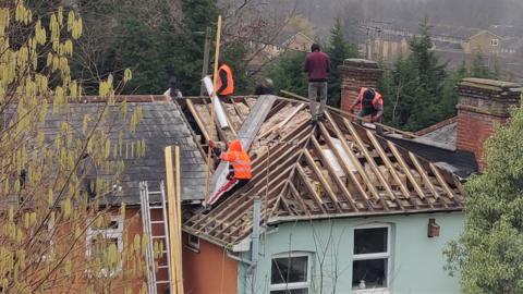 A roof of a pale blue house with workers on top of it, three of the four are wearing hi-viz clothing and the fourth is wearing a dark red jumper. A ladder is seen propped against one side of the house along with things lengths of wood