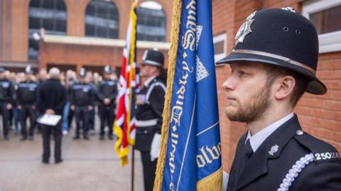 A uniformed police officer stands in the foreground, looking straight ahead. He is holding a blue and gold flag. In the background, out of focus, are a number of other police officers.