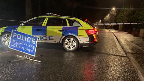 A PSNI car parked across a road with a sign in front of it saying 'Police Security Alert Road Closed'