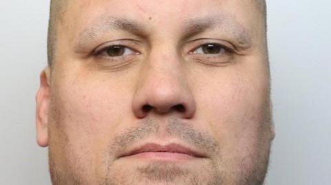 Mackay with a pale face and little facial hair, dark eyes and a shaved head looking into the camera.