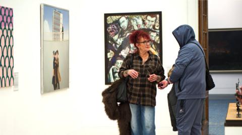 A woman with red hair tied up and glasses is wearing a chequered shirt and talking to a man in a blue hoody and tracksuit. They are standing in a gallery