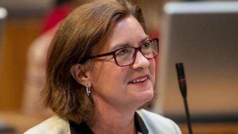 A head and shoulders picture of Eluned Morgan wearing red glasses and smiling in the Senedd chamber with a microphone in the background. She has a white jacket on, and the collars of a black shirt can be seen. She has silver earrings in.
