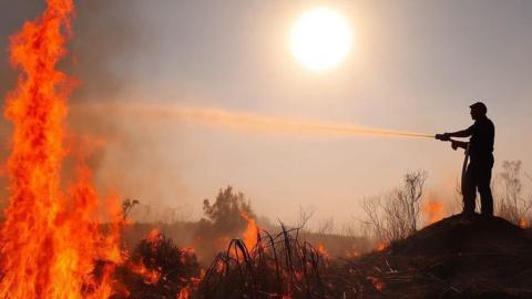 The silohouette of a firefighter tackling a live fire. He stands on a mound of dirt to the right of the image, holding a water hose, which is spraying water over gorse and hedging fully alight on the ground and a large flame rising up to the sky to the left of the image. 