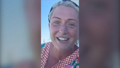 Laura Kenny smiles while speaking outside. She has blonde hair tied back with a headband, and wears a red and white checked top